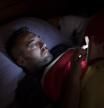 A man in glasses lies in bed, illuminated by the soft glow of his smartphone screen. He appears focused and relaxed while scrolling before sleep, wrapped in a blanket.