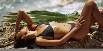 Incredibly beautiful sexy beach girl model in black swimsuit tanning on the beach and bathing in the ocean, resting on tropical rocky seashore laying with a palm leaf on the stone in the water, enjoying summer vacation