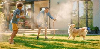 Father, Daughter, Son Play With Loyal Golden Retriever, Dog Tries to Catch Water from Garden Water Hose. Family Spending Fun Outdoors Time Together. Sunny Day Idyllic Suburban Home.