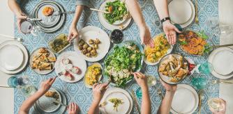 Mediterranean style dinner. Flat-lay of table with salads, starters, pastries over blue table cloth with hands holding drinks, sharing food, top view. Holiday gathering, vegetarian party concept