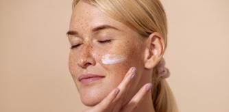 Close up of beautiful young female with cream on a face. Smiling woman with closed eyes touching cheek with fingers.