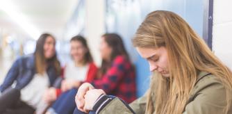 A high school girl sits alone by her locker, her head hung low in sadness, as a group of three friends in the background point at her and laugh.