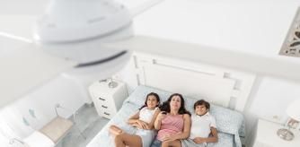 Una madre con sus dos hijos refrescándose en la cama bajo un ventilador de techo.