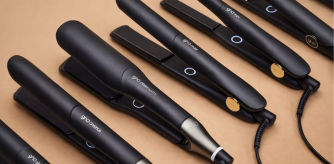 Herramientas de peinado ghd alineadas sobre fondo beige, con planchas y rizadores profesionales en color negro.