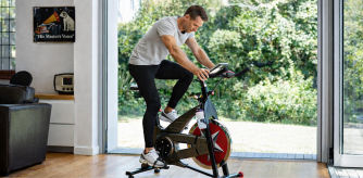 Hombre adulto haciendo ejercicio en una bicicleta estática dentro de un salón luminoso, con grandes ventanales y un jardín verde al fondo