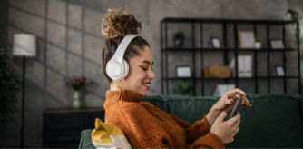 Mujer joven sentada en un sofá verde escuchando música con auriculares blancos mientras usa su móvil en un salón moderno