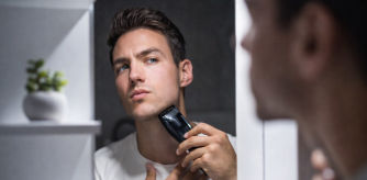 Hombre joven afeitándose el cuello con una recortadora eléctrica frente al espejo del baño, concentrado en su reflejo