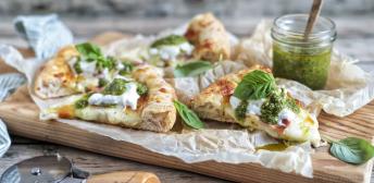 Trozos de pizza sobre una tabla de madera con un cortador y un bote de salsa pesto