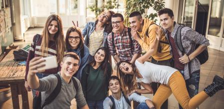 Large group of happy high school students having fun while taking a selfie with mobile phone in a hallway.
