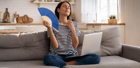Tired millennial woman suffers from stuffiness and an inoperative air conditioner, waving blue fan sitting on couch at home working on laptop computer. Overheating high temperature, hot summer weather (Tired millennial woman suffers from stuffiness an