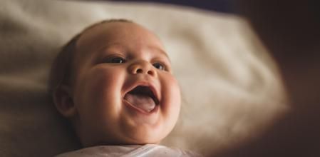 High angle view of cheerful newborn baby girl lying down on the bed and laughing while her mother, that is blurred in foreground, is playing with her.