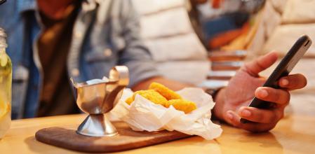Portrait of handsome successful bearded south asian, young indian freelancer in blue jeans shirt sitting in cafe with chicken nuggets and lemonade. Hold mobile phone at hand.