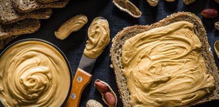 Bote de crema de cacahuete al lado de una tostada untada, un cuchillo y cacahuetes en cáscara
