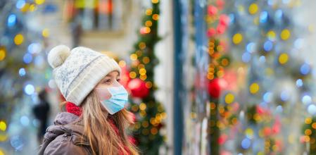 Girl wearing face mask on a Parisian street or at Christmas market looking at shop windows decorated for Christmas. Seasonal holidays during pandemic and coronavirus outbreak