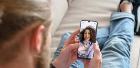 Young man sitting on sofa holding smartphone communicating with african girl friend on mobile screen, making video call using cell phone mobile social media dating app. Video call concept. Over shoulder closeup view (Young man sitting on sofa holding