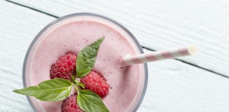 Top view of a raspberry smoothie glass set on a wooden boards, decorated with drinking straw, three raspberries and mint leaves