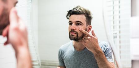 Young man taking care of his skin. Man applying skincare facial treatment cream on face in the bathroom. Handsome guy applying moisturizer and looking at himself while standing in front of the mirror.