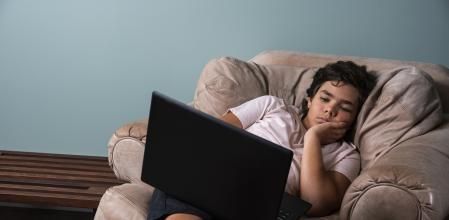 A boy reclining in a chair at home using a laptop computer. He looks tired and worn out. A concept on too much screen time and binge watching for children.