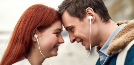 Close up of happy couple of teenagers listening to music using the same pair of earphones, looking at each other with love. Technology, romance concept. Side view. Web Banner (Close up of happy couple of teenagers listening to music using the same pai