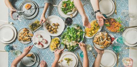 Mediterranean style dinner. Flat-lay of table with salads, starters, pastries over blue table cloth with hands holding drinks, sharing food, top view. Holiday gathering, vegetarian party concept