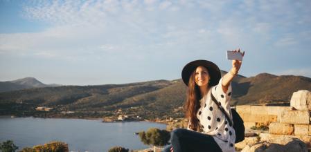 Vintage toned image of a young tourist woman, taking a selfie photo with her smartphone on the beautiful Greek seaside, standing on top of the Cape Sounion, on the most southern part of the Attica. She is wearing a casual street style outfit, a hat and a backpack, enjoying a sunny day by the Mediterranean sea, snapping some photos for her social networks or just personal collection.