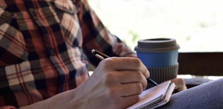 closeup of a young caucasian man wearing jeans and a plaid shirt writing with a pen in a notebook in the porch of a house or a ranch