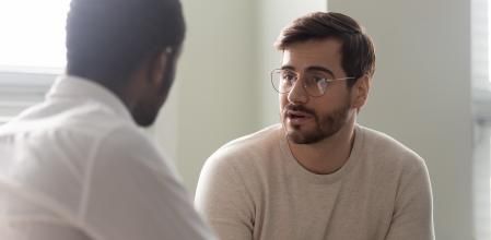 Man consulting with practitioner, African psychologist holding clipboard with card sitting in front of patient listens his mental health complaints. Job interview process applicant and HR manager concept