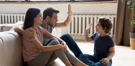 Happy young parents sit on warm wooden floor play have fun with excited preschooler son, smiling family relax rest in cozy home living room, engaged in funny game activity with little kid