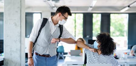 Young people with face masks back at work in office after coronavirus quarantine and lockdown, greeting.