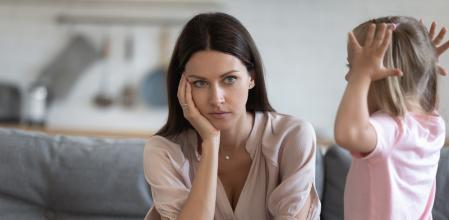 Unhappy mother having problem with noisy naughty little daughter screaming, demanding attention, child tantrum manipulation concept, tired thoughtful mum holding head, sitting on couch at home