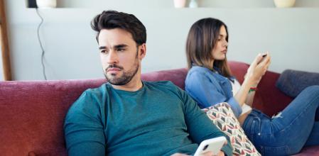 Shot of angry young couple ignoring each other using phone after an argument while sitting on sofa at home.