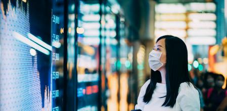 Economic and financial impact during the Covid-19 health crisis deepens. Businesswoman with protective face mask checking financial trading data on smartphone by the stock exchange market display screen board in downtown financial district showing stock market crash sell-off in red colour