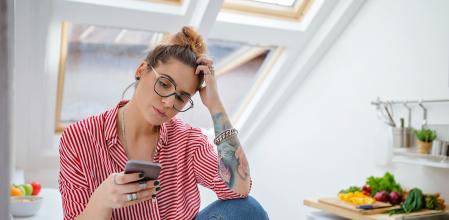 Lovely woman with eyeglasses in red and white shirt, sitting in the kitchen during breakfast using her phone.