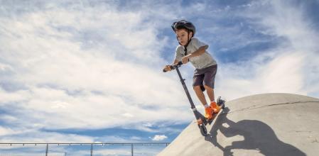 Child wearing a helmet goes down a ramp with a scooter in a skate park on a sunny summer day