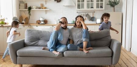 Tired mother and father sitting on couch feels annoyed exhausted while noisy little daughter and son shouting run around sofa where parents resting. Too active hyperactive kids, need repose concept