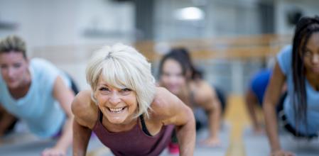 An older Caucasian woman is seen holding a plank pose while participating in a  co-ed, multi-ethnic, fitness class.  She is smiling and appearing to enjoy the class.