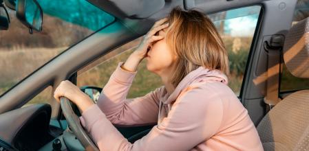 A young blonde sad woman is sitting behind the wheel of a car and holding her head in despair, covering her eyes with her hand. Side view. Stress while driving a right-hand drive car.