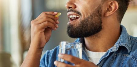 Bachelor drinking medication with a glass of water. Hispanic man drinking a supplement capsule in his apartment. Man using a glass of water to take his drugs. Sick man in recovery with a pill