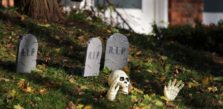 Front yard of private house decorated by fake skull, bones hands and tombstones between foliage on a green grass lawn for an old American trick-or-treat Halloween tradition.