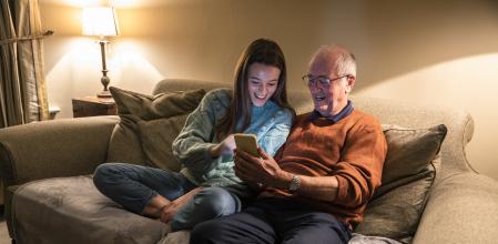 Senior man sitting with his teenage grandchild while they show him their smart phone. Both look happy and having quality time together in the warmth of the family home