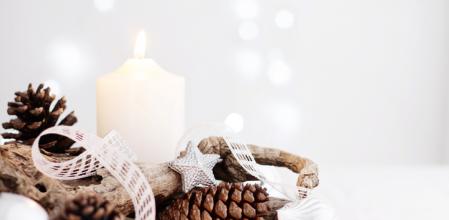 Close up of a beautiful centrepiece arranged for Christmas holidays on a table with white tablecloth. The decoration features a beautiful shaped dry branch, pine cones, white candles, silver Christmas ornaments and a delicate white bow on a silver tray. Defocused light background with Christmas lights.