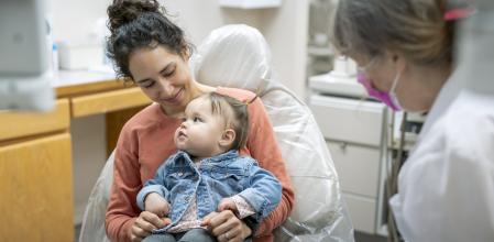 A young mixed race mother holds her calm one year old toddler in her lap during a consultation with the child's pediatric dentist.