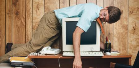 A man and office in 1980's - 1990's style, complete with vintage computer and technology of the time, sleeps on top of his desk, slumping over the computer, too tired and bored to continue working.  Wood paneling on the wall in the background.
