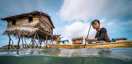 Split wide angle shot of a Bajau woman rowing wooden canoe in floating village carrying her child, showing the ocean floor and the wooden floating houses at the background, with blue sky and white clouds