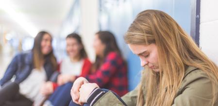 A high school girl sits alone by her locker, her head hung low in sadness, as a group of three friends in the background point at her and laugh.