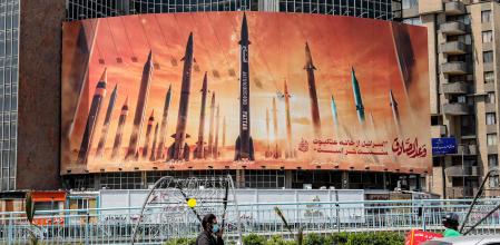 A motorcycle speaks on a phone while driving past a billboard depicting named Iranian ballistic missiles in service, with text in Arabic reading 