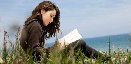 Una niña lee atentamente una novela en un rincón de vegetación junto al mar