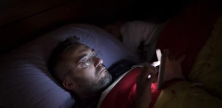 A man in glasses lies in bed, illuminated by the soft glow of his smartphone screen. He appears focused and relaxed while scrolling before sleep, wrapped in a blanket.