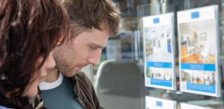 Pareja mirando el escaparate de una inmobiliaria con anuncios de venta o alquiler de casas, pisos o apartamentos.