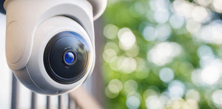 Cámara de seguridad exterior blanca con lente visible, instalada en una barandilla y fondo desenfocado de vegetación